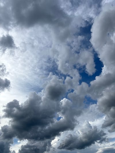 The image depicts a dramatic sky filled with a mix of dark, ominous storm clouds and brighter, wispy cirrus clouds. A patch of clear, deep blue sky is visible in the center right. In the lower portion of the image, power lines traverse the frame horizontally, indicating an urban or semi-urban environment. The overall impression is one of impending weather change. The specific location within Kurgan, Russia cannot be determined from the visual information alone.