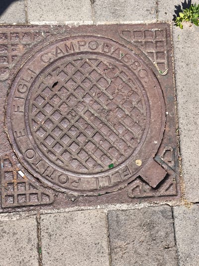 The image shows a close-up, high-angle view of a circular, cast-iron manhole cover embedded in a sidewalk composed of grey concrete pavers. The manhole cover has a raised, diamond-shaped pattern in its center and is encircled by text cast into its surface. The text appears to read "FIGLI. DI. POTTINO CAMPOBASSO." On the right side of the manhole cover, a raised, angular tab extends outwards. The edges of the manhole cover are slightly rusted and dirty. The surrounding sidewalk shows some debris, including small pieces of vegetation and dirt. In the upper right corner, a patch of green foliage is visible growing from the edge of the sidewalk. The lighting suggests it is a sunny day. The manhole cover is located in the city of Bojano, Italy, as indicated by the text "CAMPOBASSO" which is the province where Bojano is located.