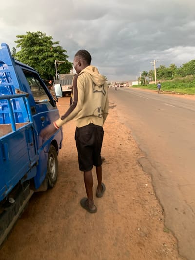 In Gbeleko, Nigeria, a young man in a light-colored hooded sweatshirt and black shorts stands next to a blue truck. He is facing away from the camera and appears to be reaching towards the truck's side. The truck is positioned on a dirt shoulder next to a paved road. The road has several vehicles in the distance, including motorbikes and trucks, suggesting a rural or semi-urban setting. The sky is overcast with heavy gray clouds, indicating impending rain or a stormy day. Lush green trees and vegetation line the side of the road. Power lines and utility poles are visible in the background. The ground is a mix of dirt and gravel.