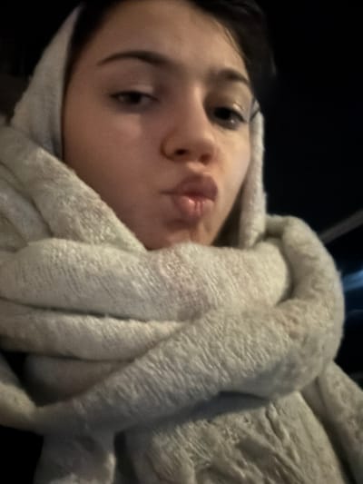 A person lies beneath a thick, light-colored blanket. The blanket wraps partially around their face, leaving their eyes, nose, and mouth visible. The person has dark hair and is angled slightly to the left. Their eyes are open and looking at the camera. Their lips are pursed slightly. The background is dark.