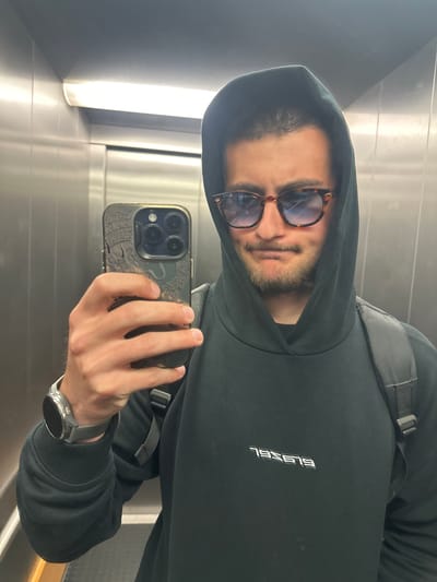 A man is depicted taking a selfie inside what appears to be an elevator, characterized by its metallic walls and vertical strip lighting. He is wearing a black hooded sweatshirt with the hood up, and a black backpack strap is visible over his right shoulder. His face is partially obscured by tortoise-shell framed sunglasses, which reflect the interior lighting. He has a short beard or stubble. The man holds a smartphone in his right hand, featuring a distinctive dark case with a dragon motif. On his left wrist, he wears a circular-faced smartwatch with a grey band. The text "ELSSEL" is visible on the front of his hoodie, which would read "LESSEL" on the actual garment due to the mirrored reflection of the selfie. The image is rotated 90 degrees counter-clockwise. The man's expression is neutral. The setting is an indoor space, likely an elevator, with the provided location context being Essen, Germany.