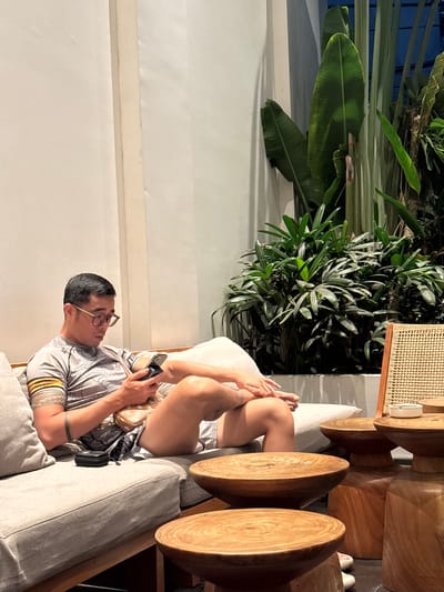 A man with glasses is lounging on a sofa, holding a phone and a small bag. He is wearing a patterned t-shirt and shorts. The sofa is light gray and situated in an outdoor setting with lush green plants, including large banana leaves, and wooden furniture. A woven rattan stool is also visible. The lighting suggests it is either late afternoon or early evening, with warm sunlight casting shadows. The scene appears to be a relaxed and comfortable outdoor living space, possibly a patio or garden area, in a location like South Quy Nhon Ward, Vietnam. There is no visible text in the image.