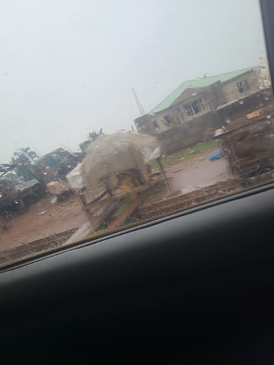 The image shows a view through a car window, with water droplets obscuring the scene outside. In the background, a building with a green roof is under construction, featuring unfinished walls and window frames. To the left of the construction site, there are more structures, some appearing to be temporary shelters or shacks. The ground is muddy and uneven, suggesting an unpaved area. Various objects, including sacks or tarpaulins, are visible, along with some scattered debris. There are no people clearly visible in the image. The overall impression is of a developing or somewhat disorganized urban environment, likely in Karu, Nigeria, given the context. The sky is overcast and hazy.