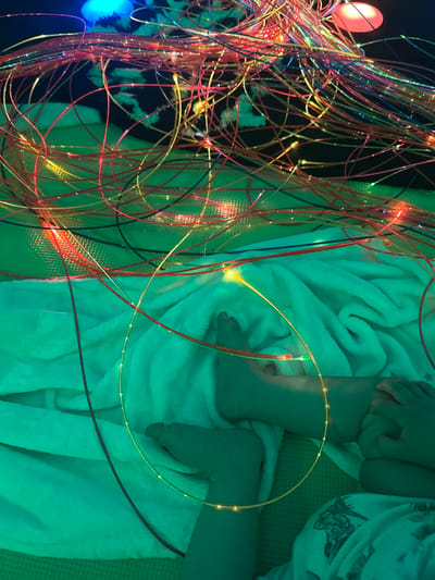 The image shows a close-up, overhead view of a baby's legs and feet, wrapped in a fluffy white blanket. The blanket is illuminated by a teal-colored light, creating a serene and calming atmosphere. Interspersed with the baby's legs are numerous thin, flexible fiber optic strands, some glowing with warm yellow and red light, others appearing to be off. One prominent fiber optic strand loops gracefully over the baby's knee and continues off-frame. The lighting is dim, suggesting an indoor setting, possibly a sensory play area or a dimly lit room designed for relaxation. The baby's limbs are visible, with one hand resting near their knee. The texture of the blanket is soft and plush. The fiber optic strands create a visually dynamic element, contrasting with the softness of the blanket and the baby's skin. The overall impression is one of gentle, immersive sensory experience.