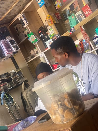 The image shows two men in what appears to be an electronics store in Barakin, Nigeria. The store is filled with shelves stocked with various electronic accessories, including phone cases, chargers, USB hubs, and what looks like phone boxes.
In the foreground, one man, wearing a grey t-shirt and a gold chain, is partially visible, looking towards the right. He is holding a clear plastic container filled with small, light brown items, possibly food or some other goods. The container has a green lid.
In the background, another man wearing a black beanie is also visible. The shelves are organized with items stacked neatly. Visible text includes brand names like "oraimo," "Redmi," "Helio G99," "Z6D," "LINNCO," and "SPORT3.0 USB HUB," suggesting a variety of electronic products are for sale.
The lighting suggests it is daytime, and the scene is indoors, within a retail environment. The overall impression is of a busy marketplace or a small electronics shop.