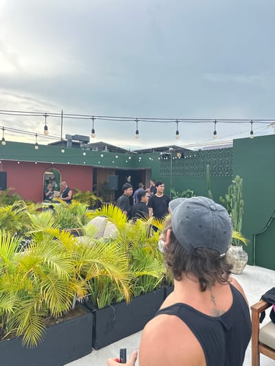 The scene depicts an outdoor rooftop event in Tibubeneng, Indonesia. A group of people are gathered near a stage or platform, possibly for a performance or presentation. The backdrop includes lush tropical foliage and cacti, with a green wall and a red building featuring an arched window. String lights are strung across the open sky, and speakers are visible on the platform. In the foreground, a person wearing a dark tank top and a baseball cap is looking towards the event, and another person with a cell phone is standing near the plants.