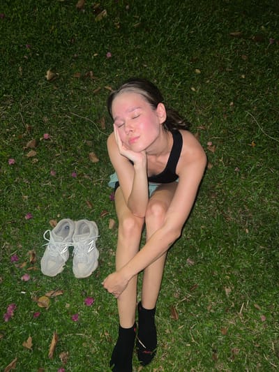 A young individual is captured in an overhead shot, lying peacefully on their side on vibrant green grass, likely in Nam Nha Trang, Vietnam. The person, appearing to be female, has dark hair with a distinctive white or blonde streak at the front, and her eyes are closed, suggesting rest or sleep. Her head is gently supported by her hands, which are crossed beneath her cheek. She wears a black athletic top or sports bra and light blue shorts, partially visible. Black ankle socks are on her feet; one sock features a red logo with white text that appears to read "Atleta."
Beside her feet, a pair of light grey New Balance sneakers with visible "N" logos are placed neatly on the grass. The ground is densely covered in green blades, interspersed with scattered brown leaves and delicate pink or purple flower petals, indicating an outdoor, natural setting. The lighting is dim and artificial, casting soft shadows, which points to the scene taking place at nighttime or late evening. The overall atmosphere conveyed is one of tranquility and relaxation.