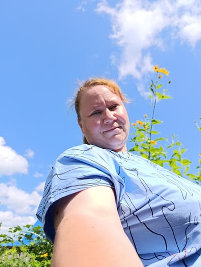The original image shows a person wearing a blue t-shirt with a black line pattern. The person has fair skin and light-colored hair. They are looking at the camera. The background consists of a bright blue sky with some white clouds and green foliage with yellow flowers. This appears to be an outdoor setting. The image was taken from a low angle, looking up at the person. No specific details about the city of Зипуново, Russia, can be inferred from the image itself.