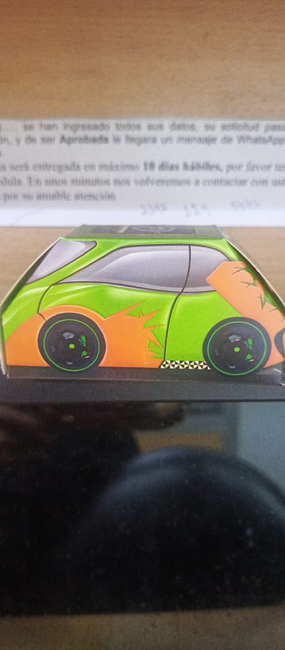 The image shows a stylized, cartoonish representation of a green and orange car, likely printed on cardboard or a similar material. The car has black wheels with green accents, and a checkered pattern runs along the bottom edge of the car. Behind the car, there is a piece of paper with printed Spanish text. The image appears to be taken indoors, with a dark object (possibly a laptop) in the foreground. The setting is not definitively established as Guayaquil, Ecuador.
