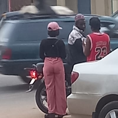 Three people are on a motorcycle next to a parked vehicle. The person in front is wearing a pink hat, a black shirt and pink trousers, and a black mask. The person in the middle is wearing a patterned top and a pink head covering. The person in the back is wearing a red basketball jersey with the number 23 and a black head covering. The vehicle parked next to them is blue. The setting appears to be urban. The location is Sardauna, Nigeria.