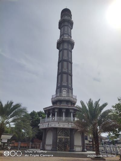 The image shows a tall, dark gray minaret with an octagonal base, topped with a dome. The minaret has multiple levels with balconies and decorative windows. Palm trees are visible in the foreground, to the left and right of the minaret. The sky is overcast with some clouds. A timestamp in the lower right corner reads "2026.06.15 10:15". A logo and text from "Vivo V15Pro AI Triple Camera" are present in the lower left corner. In the background, there are some buildings and other trees. The scene appears to be outdoors in a public space. Based on the metadata and the name of the minaret, the location is identified as Keudekruenggeukueh, Indonesia.