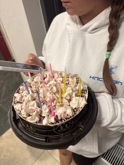 A person, likely female based on the braided hair and some visible facial features, is holding a birthday cake with lit candles and a smartphone. The person is wearing a white hoodie. The cake is round, covered in white frosting, pink sprinkles, and dark chocolate sprinkles. There are several candles on the cake, some pink, some blue, and some yellow. The person is looking down at the smartphone, which they are holding in their left hand, and the cake in their right arm. The background is blurred but appears to be an indoor setting with a light-colored wall and a tiled floor. The image does not provide any specific information or context that would allow for its location to be identified within the city of Korolyov, Russia.