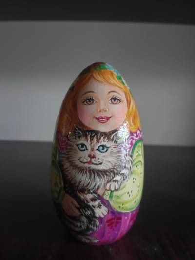 The image shows a hand-painted nesting doll with a depiction of a woman with blonde hair and a cat. The doll is an oval shape and is intricately decorated with colors and patterns. The woman has large eyes, rosy cheeks, and red lips. She is holding a fluffy tabby cat with blue eyes. The cat is positioned in front of the woman's torso, appearing to be cradled in her arms. The background is blurred, showing a neutral gray wall and a dark surface below the doll. There is no information in the image to suggest a location in the city of Kherson, Ukraine.