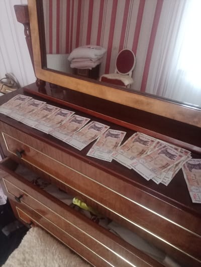 A dresser with a mirror is in the center of the frame. The dresser is made of polished wood with gold trim and has three drawers. The top drawer is slightly ajar, revealing some contents within. On the surface of the dresser, to the left, a stack of Moldovan leu banknotes is laid out. These banknotes appear to be the same denomination, with the number "200" visible on some of them. The mirror reflects a portion of a room, showing striped wallpaper in shades of pink and white, a radiator, and a small red chair with a white fluffy cushion. To the right of the chair, a white curtain is visible. The overall scene suggests an indoor setting, likely a bedroom or living area.