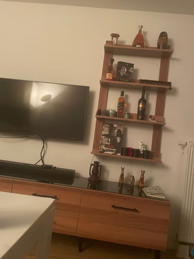 The image depicts an indoor scene, likely a living room, in Yunus Emre, Turkey. A large, dark-screen television is mounted on a light-colored wall to the left of the frame. Below the television, a black soundbar is positioned on top of a wooden media console with a dark glass top. The media console features several drawers with dark handles.
To the right of the television, a multi-tiered wooden shelf unit is mounted on the wall. The shelves are adorned with various decorative items, including bottles of alcohol, books, and small figurines. Notable items on the shelves include an hourglass, a bottle labeled "Mone," a dark brown decorative vase, a box with an image of Mustafa Kemal Atatürk, a bottle of Johnnie Walker Black Label, another dark bottle, a small lantern, and several cat figurines and an Egyptian pharaoh figurine on the media console. A white teddy bear and two black cylindrical containers are also visible on one of the shelves. A spiral-bound notebook rests on the media console.
The overall environment appears to be a residential space with a modern aesthetic, characterized by the combination of the wooden furniture and the minimalist wall-mounted shelves. The lighting is dim, suggesting an evening or indoor setting.