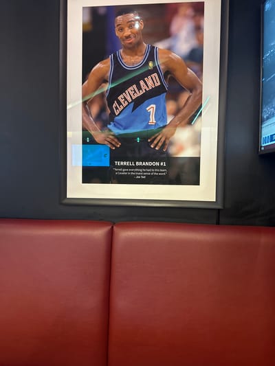 Here is a forensic description of the image:

The image depicts a framed poster of Terrell Brandon, a former basketball player for the Cleveland Cavaliers. The poster is mounted on a dark gray wall panel next to a red, padded seating area, commonly found in public places. The poster features Brandon wearing a Cavaliers jersey with the number 1. Text accompanying the image credits a quote to Joe Tait, a famous Cleveland announcer. Above the poster, a television screen is mounted on the wall, though the content is out of focus. The scene appears to be indoors, likely inside an establishment in Cleveland, Ohio.