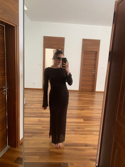 In this mirror selfie, a woman stands in what appears to be a modern interior with white walls and wooden floors and accents. She is wearing a long-sleeved, dark brown, floor-length dress with a textured knit pattern. She has dark hair styled in a bun and is wearing sunglasses. She is holding a smartphone in her right hand, which she is using to take the photo. Her left hand is resting on her hip. Her bare feet are visible at the bottom of the dress. The reflection shows a doorway with wooden paneling and a metal handle in the foreground. Behind her, also reflected in the mirror, are two doors with wooden panels and a wooden accent above them. The lighting suggests it is daytime. The scene is clean and well-lit, indicative of a residential interior.