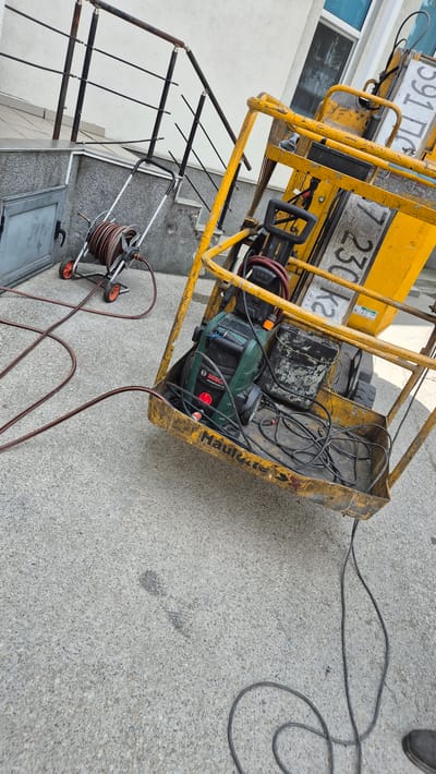 An outdoor scene in Sevlievo, Bulgaria, features a yellow aerial work platform with a basket containing a green Bosch pressure washer, a black toolbox, and various electrical cords. A red hose is connected to the pressure washer and extends out of the basket. A hose reel with a red hose is visible on the left side of the image, near a grey metal door and a set of stairs with a metal railing. The ground is paved with concrete. The scene appears to be set up for work or cleaning.