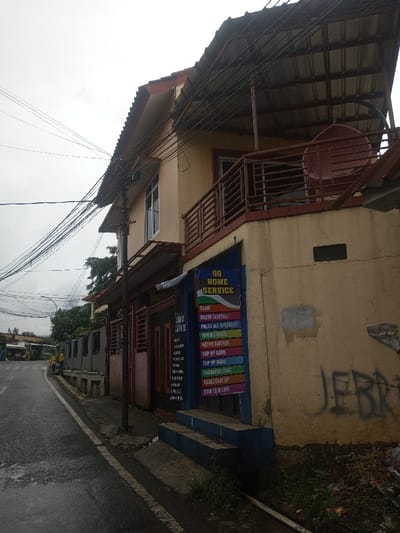 A two-story building with a rusty metal roof and balcony is shown next to a wet road. A sign reads "QQ HOME SERVICE" with a list of services including "SEDUR, MUOER INTERNET, PULSA ALL OPERATOR, TOKEN LISTRIK, BAYAR TAGIHAN, TOP UP GAME, TOP UP DANA, TRANSFER UANG, AKSESORIS HP, DAN LAIN LAIN." Graffiti is scribbled on the side of the building, and power lines run overhead. A red hanging chair is on the balcony.