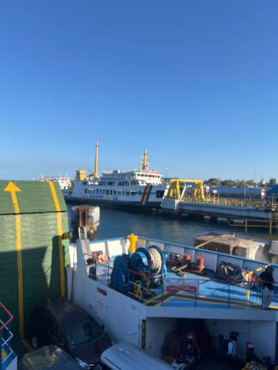 The image shows a ferry docked at a port. The ferry is white with blue and yellow accents. Several cars are parked on the ferry's deck, including a dark colored sedan and a silver car. There are also people on the ferry, some of whom appear to be passengers. In the background, another ferry is docked, and there is a yellow structure that appears to be part of the port's infrastructure. The sky is clear and blue. The location is the port of Ketapang, Indonesia, which is a major ferry terminal connecting the island of Java to the island of Borneo.