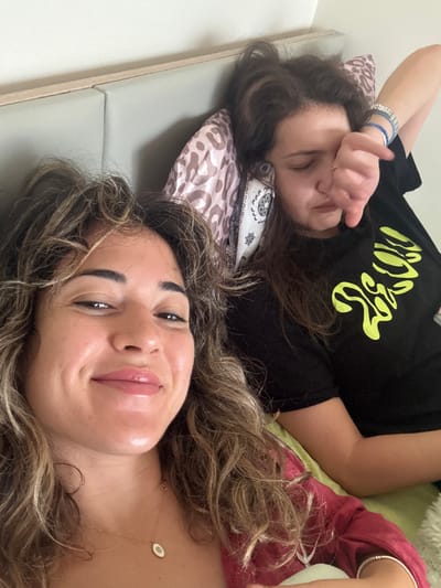 This image depicts two females indoors, presumed to be in a bedroom setting, located within the city of Kalyvia Thorikou Municipal Unit, Greece.
The subject in the foreground, positioned on the left, is a female with long, wavy brown hair featuring lighter highlights. She is looking directly at the camera with a slight smile and glossy lips. She wears a dark red garment and a delicate gold chain necklace with a small white oval pendant.
The second subject, positioned behind and to the right of the first, is also a female with long, dark brown, wavy hair, appearing somewhat disheveled. She is lying down, resting her head on a pillow with a light brown and white abstract pattern. Her left hand is raised, with fingers partially obscuring her left eye and nose. Her facial expression appears neutral or slightly strained. She is wearing a black t-shirt with a bright yellow/lime green abstract graphic design on the chest and multiple blue and silver bracelets on her right wrist.
The immediate environment includes a light-colored padded headboard with a visible wooden trim along the top. A plain, light-colored wall is visible above the headboard. A light green fabric, possibly a blanket or additional pillow, is visible beneath the second subject's right arm. The overall lighting is bright and even. The action suggests the foreground subject is taking a selfie, while the background subject is in a state of rest or disengagement from the camera.