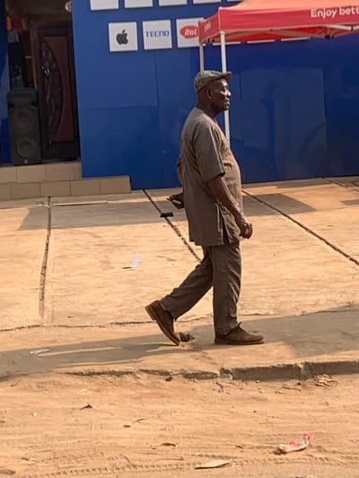 An adult male is observed walking from right to left on a concrete sidewalk in Aguleri, Nigeria, adjacent to a dirt road. He wears a light brown tunic, matching trousers, a patterned cap, and dark brown shoes, and carries a black object in his left hand. His head is turned to the right, facing away from the direction of travel. The sidewalk shows cracks and leads to a dusty dirt ground with tire tracks and a discarded red and white plastic item in the foreground. In the background, a building with a royal blue facade displays brand logos for Apple, TECNO, and itel. To the left of the man, a dark speaker stands next to a wooden door. A red canopy with partially visible text "Enjoy bett" is present on the far right. The scene is illuminated by strong direct sunlight, casting distinct shadows.