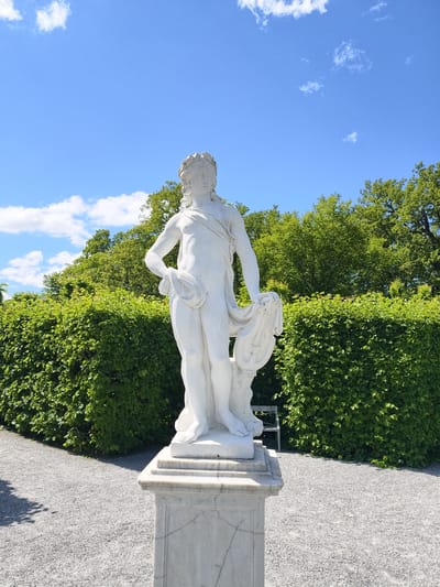 A white marble statue of a male figure wearing a laurel wreath stands on a marble pedestal in a well-maintained garden. The statue depicts the figure with a cloth draped over its left arm and shoulder, and the right hand resting on its hip. Behind the statue, a meticulously trimmed green hedge forms a backdrop, with lush green trees visible beyond the hedge. The ground in front of the statue is covered with fine grey gravel. In the distance, a park bench can be seen peeking out from behind the hedge. The sky above is a bright blue, dotted with fluffy white clouds. The scene is bathed in natural daylight, suggesting it is a clear, sunny day. This location appears to be the Drottningholm Palace Park, situated in Ekerö kommun, Sweden, known for its baroque gardens and classical sculptures.