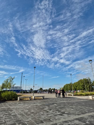 An outdoor scene captures a bright, clear blue sky filled with wispy, streaky white clouds, possibly cirrus or cirrocumulus. The foreground features an expansive paved walkway or plaza made of grey, rectangular block paving stones, showing distinct shadows stretching across its surface from the left, indicating either early morning or late afternoon sunlight.

Along the left side of the paved area, several people are walking casually. In the mid-ground, a group of three individuals is visible, with other figures further in the distance. Modern streetlights with distinctive D-shaped or circular light fixtures are regularly spaced along the walkway, casting long shadows. Concrete benches or planters are also integrated into the paved design.

Beyond the walkway, a line of lush green trees and bushes forms a natural barrier. Peeking through the trees in the distance are several parked cars and a blue industrial-style building. Text visible on the side of this building reads "AngliaCable." To the far left, a tall, lattice-work structure, possibly a communications tower or mast, rises against the sky. The overall impression is a spacious, well-maintained public area on a sunny day in West Suffolk, United Kingdom.