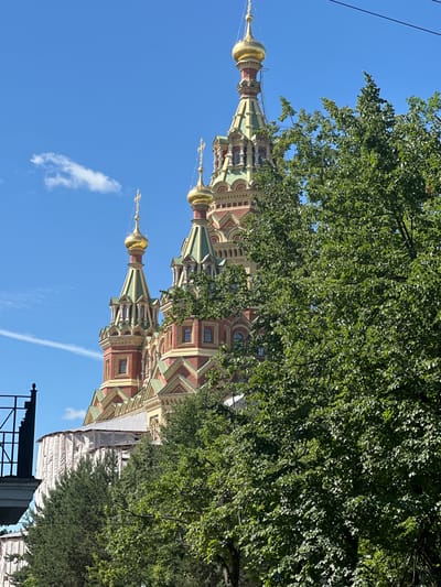 This is a vertical, eye-level, medium shot photograph of the Peterhof Grand Cathedral (also known as the Cathedral of Saints Peter and Paul) in Peterhof, Russia, on a clear, sunny day with a bright blue sky. The cathedral's prominent feature is its onion domes, gilded with gold, which are visible above a dense canopy of green trees. The cathedral's architecture includes red brickwork with yellow and green decorative elements. Several crosses top the domes. Some sections of the cathedral appear to be under renovation, covered in white tarpaulin. A black metal railing and part of a building are visible in the lower left corner, partially obscured by trees. The overall scene captures the grandeur of the cathedral amidst lush, green foliage.