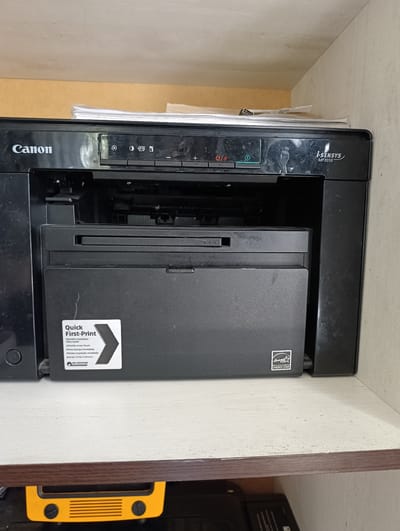 The image shows a black Canon i-SENSYS MF3010 printer situated on a shelf. The printer is black and appears to be made of plastic. There are several buttons and indicator lights on the top panel of the printer. A stack of papers is visible on top of the printer, peeking out from the front. A label with the words "Quick First-Print" is affixed to the front of the printer, along with an Energy Star logo. The printer is positioned on a white shelf with a wooden edge. Below the shelf, a yellow object with black knobs and a grille is partially visible, along with some black cables. The background consists of a light-colored wall and a wood-paneled surface above the shelf. The overall environment appears to be an indoor setting, possibly a home office or storage area. There are no people or animals present in the image. The specific location of Leñgir, Kazakhstan, is not indicated by any visible features within the image.