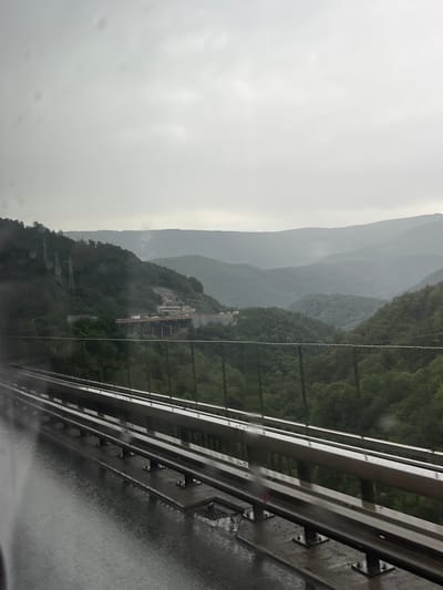 The image depicts a cloudy, overcast day, suggesting it is either early morning or late afternoon. The foreground features a metal guardrail, likely from a highway or elevated walkway, with a wet, reflective surface indicating recent rain or ongoing precipitation. Beyond the guardrail, a lush, green mountainous landscape unfolds. The mountains are covered in dense forestation, with several layers of peaks receding into the distance. In the mid-ground, nestled on the mountainside, is a complex of structures that appear to be under construction or a quarry site, with visible machinery and unfinished buildings. The overall atmosphere is somber and somewhat dramatic due to the weather and the scale of the natural and man-made features. There are no visible people or text in the image.