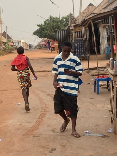 A man in a blue and white striped shirt and dark shorts looks at his phone while standing on a dirt street. Next to him, a person in a red shirt and patterned skirt holds a plastic bottle. Both are wearing sandals. The street has tire tracks and litter. Buildings line one side of the street, including a shop with chairs displayed. A few people and vehicles are seen further down the road. The sky is overcast.
