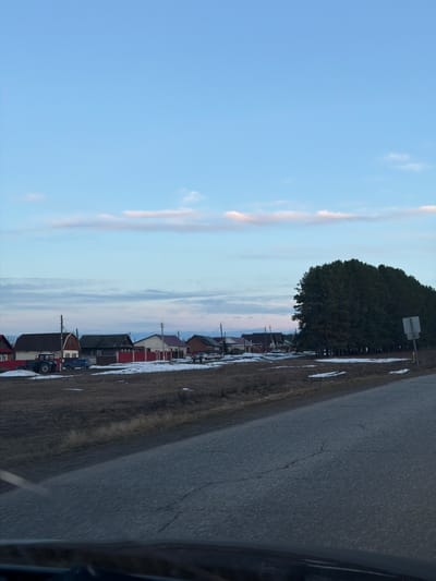 The image depicts a view from a vehicle traveling along a road in or near Дулесово, Russia. To the left is a clear blue sky with thin, wispy clouds. Along the right side of the image are houses and buildings of various colors and styles, interspersed with trees and patches of snow. A large, dark green tree stands prominently near a sign. The road is asphalt, with dry grass on either side and patches of snow. The asphalt road is cracked and dry.