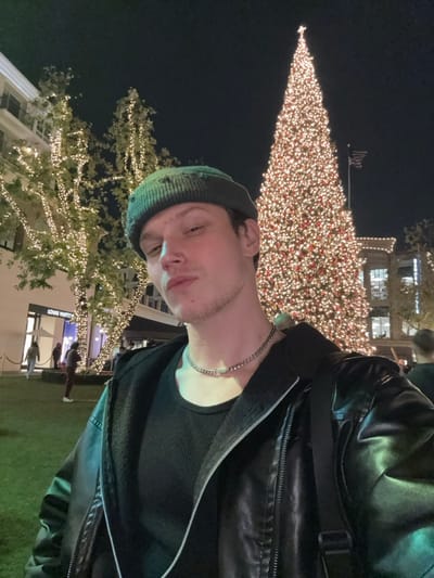 A man with a beanie, chain necklace, and leather jacket poses with a Christmas tree and lights in the background. The tree is decorated with many small lights. A building with lit windows is visible behind the tree. Another tree with white lights stands to the side, with a Louis Vuitton store underneath. People are blurred in the background.