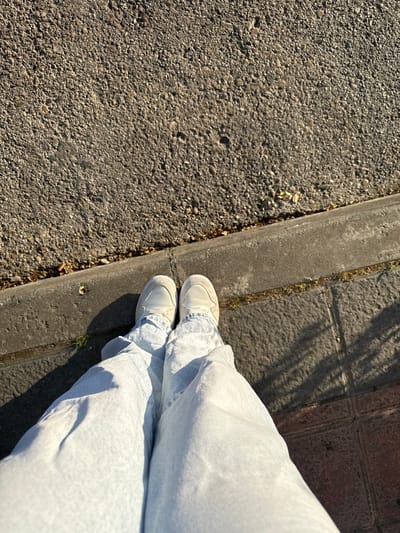 An overhead perspective shows a person's legs and feet, clad in light blue, loose-fitting jeans and white sneakers. They are standing on a tiled pedestrian area next to a concrete curb. Beyond the curb, a grey asphalt surface with visible aggregate forms the background. The scene is illuminated by the low-angled sunlight of what appears to be late afternoon, casting shadows across the tiles. A few sparse green shoots of grass are visible along the edge of the curb, and some fallen leaves are scattered on the asphalt. The image conveys a sense of an ordinary urban setting in Getafe, Spain, possibly a park or a sidewalk.