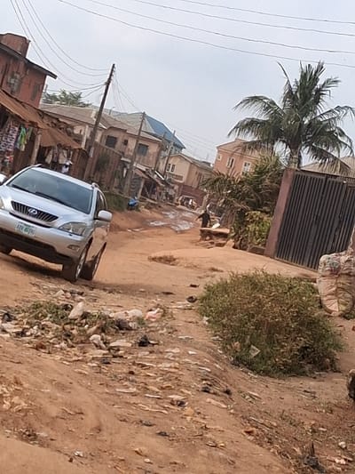 A street scene in Egan, Nigeria, features an unpaved, reddish-brown dirt road under an overcast sky. The foreground is marked by significant litter, including plastic and paper debris. A silver Lexus RX SUV, with license plate KFV 483NS, is positioned on the left side of the road. To the right, a dense green shrub and stacked white sacks are visible. The midground road exhibits muddy depressions and extends towards various multi-story buildings. On the left, a reddish-brown structure with a corrugated metal awning displays hanging garments from a market stall, adjacent to a partially visible individual in a white garment. On the right, a dark metal gate precedes a tall palm tree. Another individual, dressed in dark attire, walks away from the viewer on the right side of the road. Further down the street, a tricycle (keke napep) is discernable among additional residential and commercial structures. Overhead utility lines are suspended between wooden poles across the scene.