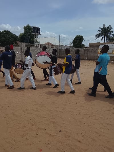 The image shows a group of young men marching in a parade, likely a school band, in an open sandy area. They are carrying drums and other musical instruments. The men are dressed in white pants, white shirts, and blue vests with a yellow collar, except one man wearing a blue shirt. They are marching in a single file line. The scene is located outdoors in a sandy area with a low brick wall and a tall water tank in the background. There is a metal coffin in the background, suggesting a funeral procession. The sky is blue with some clouds. The location provided is not in Mukaiya, Nigeria, as those coordinates correspond to a location in the Atlantic Ocean. The location is likely a small town or village in Nigeria.