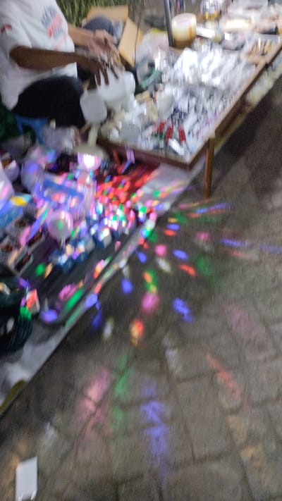 The image depicts a street scene, possibly a market stall, in Sukodono, Indonesia. A person wearing a white t-shirt and dark pants is visible, their hands engaged in manipulating white objects on a table. The table is laden with various items, many of which appear to be clear or white plastic containers, possibly for sale. Colorful lights, likely from a disco ball or similar lighting effect, are projected onto the table and the surrounding ground, creating a vibrant, abstract pattern of red, blue, green, and pink hues. The ground appears to be paved with textured material, possibly stone or concrete. In the background, greenery is visible, suggesting an outdoor setting. The overall impression is of a lively, perhaps evening or night, market or event.