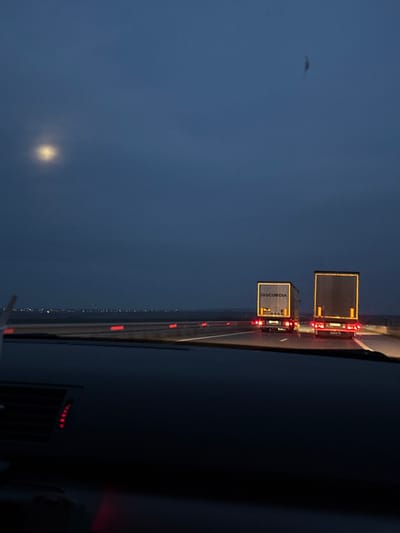 The image captures a nighttime scene from within a vehicle traveling on a multi-lane highway in the vicinity of Dobri Dol, Bulgaria. The lower portion of the frame reveals the dark dashboard and a segment of the windshield, with an illuminated red vent visible on the dashboard's left side. Ahead, the wet asphalt road features clear white lane markings. Two semi-trailer trucks are observed driving in the same direction. The truck positioned on the left has "DISCORDIA" clearly visible on its rear trailer. Both trucks display active red taillights and prominent yellow/amber reflective striping on their trailers. A median barrier to the left of the road is marked with multiple red reflectors. On the distant horizon to the left, diffuse lights, consistent with an urban or industrial area, are perceptible. The sky overhead is dark blue-grey and appears overcast, with a bright, luminous moon positioned in the upper left quadrant. A small dark smudge or object is present in the upper right portion of the sky. No individuals are visible within the frame.