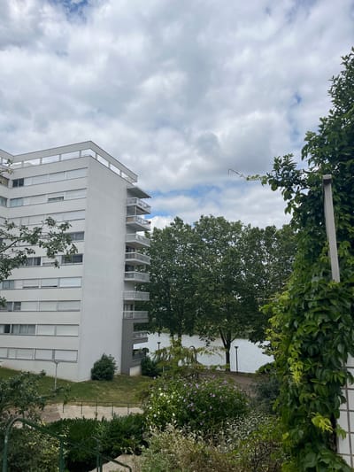The image depicts a view of a residential building and its surroundings in Créteil, France. The building is a modern, white, multi-story apartment complex with balconies on one side. Large trees with dense green foliage are visible in the middle ground, with a body of water, likely a lake or river, behind them. The foreground features various plants and greenery, including flowering bushes and climbing ivy on a white tiled structure. The sky is overcast with a mix of white and gray clouds. The scene appears to be a residential area with green spaces and water features, characteristic of certain neighborhoods in Créteil, such as those near the Lac de Créteil. There are no people visible in the image.
