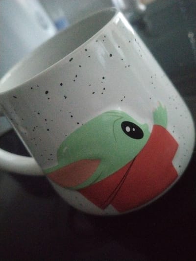 A white ceramic mug with a speckled black pattern lies tilted against a dark, indistinct background. The mug features a prominent illustration of a green, large-eyed cartoon character, widely recognized as Grogu (Baby Yoda) from The Mandalorian. The character is depicted wearing a red Santa hat, suggesting a holiday theme. Its large black eyes have a small white highlight, and a subtle smile is visible on its face, conveying a cute and endearing expression. A faint reddish hue is present on the character's ear, matching the red hat. The background is mostly dark and out of focus, with some lighter, blurry reflections suggesting an indoor setting on a reflective surface. No specific activity is taking place; the image is a static portrait of the mug. There is no visible text on the mug or in the scene. The image was captured in Crauthem, Luxembourg.