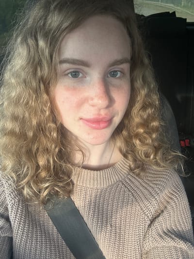 A selfie of a light-skinned person with curly blonde hair and light blue eyes wearing a beige knitted sweater and a gray seatbelt. The background appears dark, suggesting the interior of a vehicle. The location in the city of Vasilyevo, Russia, is undetermined from the image.