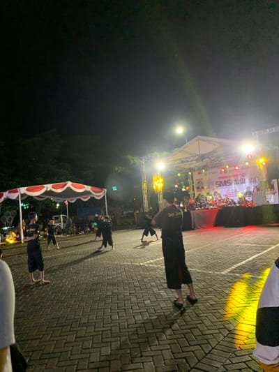 An outdoor nighttime scene reveals a stage with bright lights and a crowd. Several people are standing on a brick-paved ground, wearing dark clothes. The image is taken from a slightly elevated angle, showcasing the event's setting and the participants present. A canopy-like structure with white and red draping is also visible on the left side of the frame.