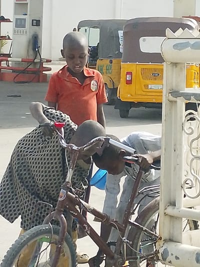 The scene depicts a petrol station forecourt in Babban Giji, Nigeria, under bright daylight. Three young individuals are present. On the left, a petrol pump station with two gray pumps and red hoses is visible against a white building. In the mid-ground, two yellow motor tricycles (Keke Napep-style) are parked.

In the foreground, center, a boy with a shaved head and an orange short-sleeved polo shirt stands upright, looking downwards with his mouth slightly ajar. To his left, partially obscured, another individual, also with a shaved head, is bent over a dark brown bicycle, wearing a dark patterned garment. This individual holds the bicycle's frame near the handlebars. To the right of the first individual, a third individual, with a shaved head and wearing a light-colored striped long-sleeved shirt, is also bent over, interacting with the handlebars of a bicycle. A small blue bucket or container is visible on the ground near these two individuals and the bicycles. On the far right, a white painted metal fence or gate with decorative scrollwork is partially visible. The ground is paved.