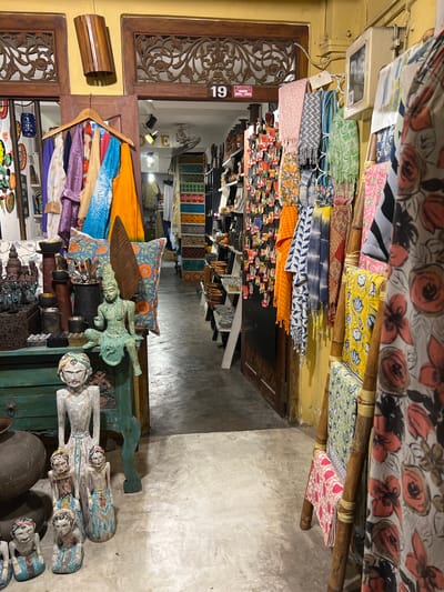 This is a vertically oriented, eye-level, medium shot of a shop interior in Galle, Sri Lanka. The shop appears to be selling textiles and handcrafted items. In the foreground, on the right side of the image, there are several wooden figures of people with painted faces, some stacked atop one another on a dark green cabinet. To the left of these figures is a blue and orange patterned pillow. A bronze-colored winged figure rests on top of a cylindrical object, with a textured leaf-like object behind it.
In the middle ground, bolts of colorful fabric are displayed vertically, some with floral patterns and others with geometric designs, suspended by wooden poles. A rack of folded textiles, in various colors and patterns including orange, blue, purple, and yellow, is visible. A wooden hanger is placed on a rack of clothing. In the background, shelves are stocked with more textiles and decorative items.
The lighting suggests it is daytime, with artificial light illuminating the interior. The scene appears to be a bustling marketplace or a souvenir shop, typical of a tourist location. Visible text includes the number "19" on a sign near the top left of the image. The overall impression is one of vibrant colors and a rich array of artisanal goods.