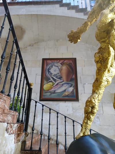 The image shows a still life painting of fish, lemons, a pitcher, and a shell, displayed on a stone wall. To the left of the painting, a marble staircase with black wrought-iron railings ascends. To the right, the lower part of a large, textured gold sculpture of a human figure is visible. The scene is indoors, likely in a public space like a museum or cultural center, given the presence of art and architectural features. The context provided indicates this is the Museum of Contemporary Art in Alicante, Spain.