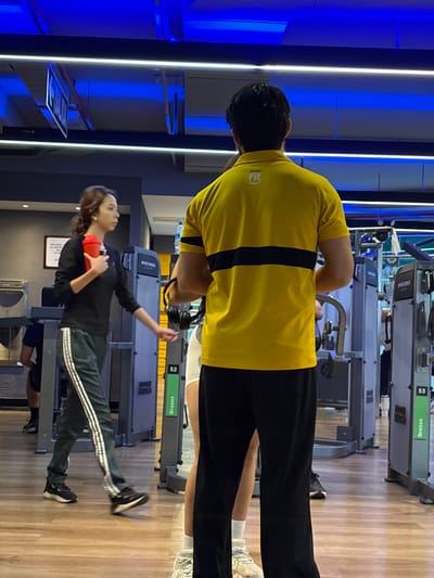 The image displays a modern gym interior, rotated 90 degrees clockwise from its natural orientation. In the foreground (right side when correctly oriented), a man is seen from the back, wearing a bright yellow short-sleeved shirt with a black horizontal stripe and black pants. His shirt features a visible "Smart Fit" logo. Headphones are around his neck. He appears to be observing or waiting, with another person's lower legs and white sneakers partially visible beside him.
In the mid-ground (left side when correctly oriented), a woman stands near gym equipment. She is wearing a black long-sleeved top and dark track pants with white stripes down the sides. She holds a red shaker bottle in her hands and appears to be looking towards the front of the gym. Her hair is brown. Behind her, a small framed sign on the wall is visible, with text that appears to read "PROHIBIDO FUMAR. LA GERENCIA." (No smoking. Management).
The gym is equipped with various machines, including those labeled "HAMMER STRENGTH" and "Brazos," with numbers like "52" and "53" visible on some. The floor is made of light-colored wood laminate. The walls are adorned with vertical LED strip lights, some emitting bright white light and others a strong blue, creating a contemporary and well-lit atmosphere. Parts of other gym users' legs are also visible in the background, suggesting an active environment. The setting appears to be a fitness center in San Isidro, Peru, during an indeterminate time of day, relying on artificial lighting.