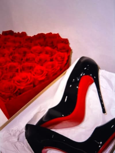 A heart-shaped box filled with red roses is positioned to the left of a pair of black high-heeled shoes with red soles, resting on white tissue paper within a shoebox. The background is a plain, light-colored surface. There are no people or any indication of the city of Beșalma, Moldova.