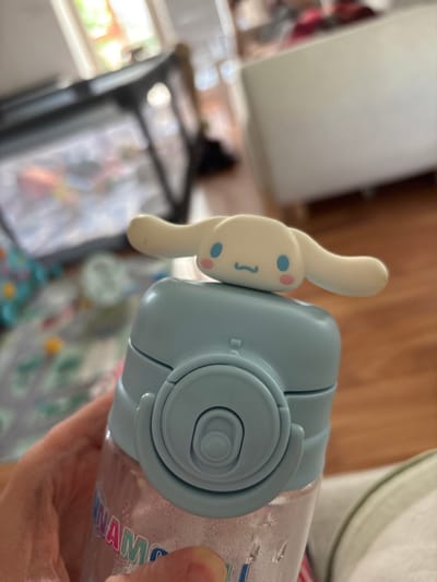 A person's hand is holding a light blue water bottle with a white cartoon character attached to the lid. The character has two blue dots for eyes and pink circles for cheeks, with a smiling mouth. The water bottle has a ridged lid and some condensation on its clear plastic body. Text on the bottle reads "INAMO". The background is blurred, showing what appears to be an indoor setting with wooden furniture and a soft, diffused light, suggesting daytime. The scene is focused on the water bottle and the hand holding it.