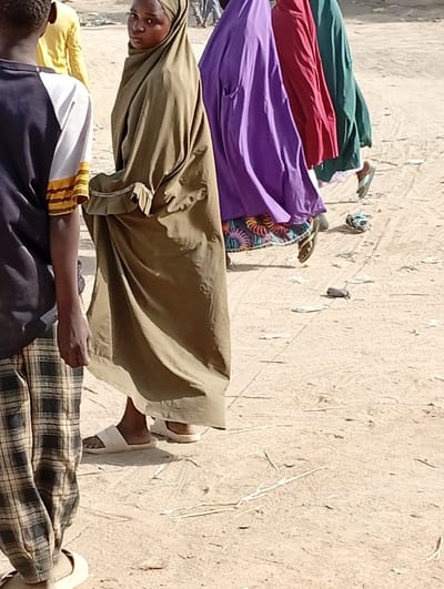 The image depicts a dusty, unpaved outdoor environment in Gayawa, Nigeria, under bright daylight conditions. Five individuals are present. In the left foreground, the back and left side of a person wearing a dark and white short-sleeved top with a yellow stripe, and brown and black plaid trousers, are visible. This person wears light-colored slip-on shoes. To their right, a young female, dressed in a full olive-green hijab/khimar with a ruffled trim near the waist, looks directly at the camera, slightly over her left shoulder. She wears white sandals. Behind her, a line of three additional individuals walk away from the camera. The first in this line wears a purple full-body covering over a patterned dress and flip-flops. The second wears a red full-body covering, and the third wears a dark green full-body covering. Scattered debris, including small white fragments and dried grass, is present on the ground. Vehicle tracks are discernible in the dust.